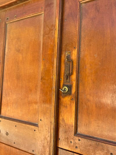 A close-up, angled view depicts a weathered, reddish-brown wooden double door, reportedly located in Φάρος, Greece. The door features paneled construction with visible wood grain and small, dark fasteners along its frame. On the right door leaf, an aged, dark-colored, surface-mounted lock mechanism is present. This mechanism consists of a vertical body with a horizontal, U-shaped pull handle at its upper section and a keyhole escutcheon below. A single, brass-toned key is inserted into the keyhole, turned approximately 45 degrees clockwise. The wood surfaces exhibit signs of wear, including scratches, dust accumulation, and variations in color saturation consistent with weathering. No individuals or active interactions are depicted; the scene is solely focused on the architectural detail of the door.