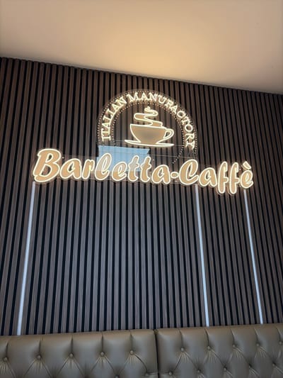 A neon sign reading "Barletta-Caffè" in a cursive font with a beige glow is mounted on a dark wooden slatted wall. Above the text, a circular neon sign displays "ITALIAN MANUFACTORY" in a circular arrangement of dots and letters, with a stylized coffee cup in the center. Two vertical white LED light strips are embedded in the wall on either side of the sign. Below the wall, a tufted leather sofa is visible. The image is taken from a frontal perspective at eye level. The setting is likely an interior space, possibly a cafe or restaurant, given the signage. There are no people or discernible actions depicted.
