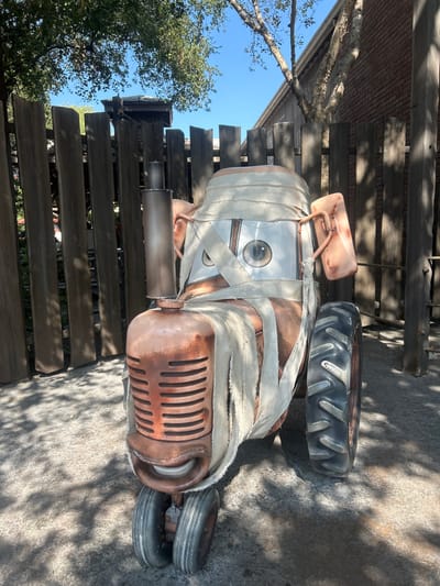 The image depicts a static, life-sized prop of a rust-colored tractor, designed to resemble the anthropomorphic tractors from Disney Pixar's "Cars" franchise, specifically adorned as a mummy. The tractor's body is extensively wrapped in numerous strips of off-white, tattered fabric. Its "face" features a rectangular white panel with large, painted, realistic eyes, and a grille below suggests a smiling mouth. A dark gray vertical exhaust pipe extends upwards from the left side of the engine block. Two small front wheels and one large, black-treaded rear wheel are visible. The prop rests on a light gray paved surface, dappled with dark shadows from overhead foliage. In the background, a tall, weathered wooden picket fence stands, behind which a building with reddish-brown brickwork and a light-colored wooden roofline is partially visible. Lush green tree foliage and branches are prominent against a clear blue sky. The scene is outdoors under bright daylight. This display is located in the Cars Land area of Disney California Adventure Park in Anaheim, California, United States.