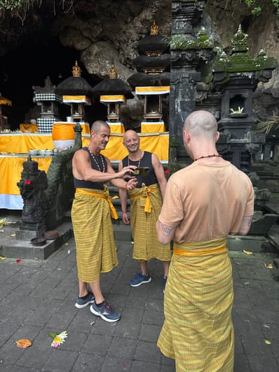 The image depicts three bald men in an outdoor setting with ornate stone structures and vibrant yellow fabric. The man on the left, wearing a black tank top and a yellow sarong, is holding a dark object, possibly a phone, and appears to be showing it to the man in the center. The man in the center, also in a black tank top and yellow sarong, is smiling and looking at the object. The man on the right, facing away from the camera, wears a light brown t-shirt and a yellow sash, and has a tattoo on his forearm. Behind them, there are several tiered structures adorned with dark roofs and golden spires, draped with yellow cloth and white and black checkered fabric. A stone serpent statue is visible on the left. The ground is paved with grey stone tiles. The background features a dark cave entrance and lush greenery overhead. This scene is located at Goa Gajah (Elephant Cave) in Dawan, Indonesia, a significant archaeological site and tourist attraction.