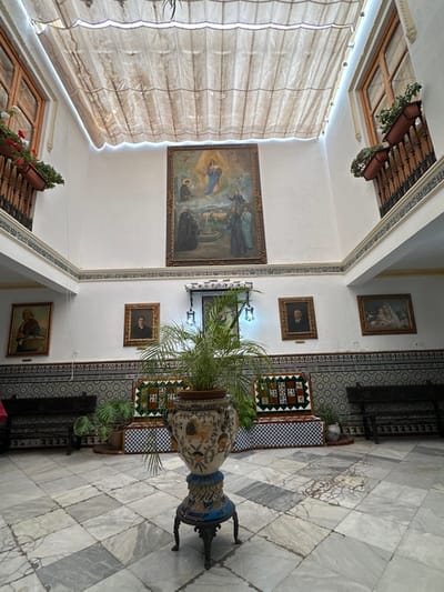 This image captures the interior courtyard of a building in Ronda, Spain. The scene is devoid of human presence. A high-angle, wide-shot perspective frames a multi-story, open-top space with a pleated, translucent fabric canopy stretched overhead, providing diffused light. The canopy shows some signs of staining and wear, with white strip lighting visible along its edges.

The walls feature a white upper section and a lower wainscoting comprised of intricate blue, white, and brown patterned ceramic tiles (azulejos). Two wooden-framed windows, each with a small dark metal balcony railing and flower boxes containing red and green plants, are visible on the upper level of the left and right walls.

A large, framed religious painting depicting the Virgin Mary and child in the sky above a landscape with multiple robed figures is centrally affixed to the back wall. Below and to the left of this painting, two ornamental lamps with multiple light sources hang from a decorative bracket. Flanking the central religious painting are several smaller, dark-framed portraits: a man in a dark suit to the right, and an older woman and another individual in dark attire on the left wall. A smaller religious painting, possibly depicting the Madonna and child, is visible on the far right wall.

The floor is composed of a mosaic of square marble or stone tiles in shades of white, grey, and light brown, exhibiting natural veining and some surface cracks. In the foreground, slightly off-center, stands a large, ornate ceramic vase painted with intricate designs, including what appear to be animal figures. This vase rests on a dark, decorative metal tripod stand and contains a vibrant green, multi-fronded potted plant. Dark carved wooden benches are positioned against the tiled walls on the left and right sides, beneath the smaller portraits. Additional small potted plants are visible at the base of the walls.
