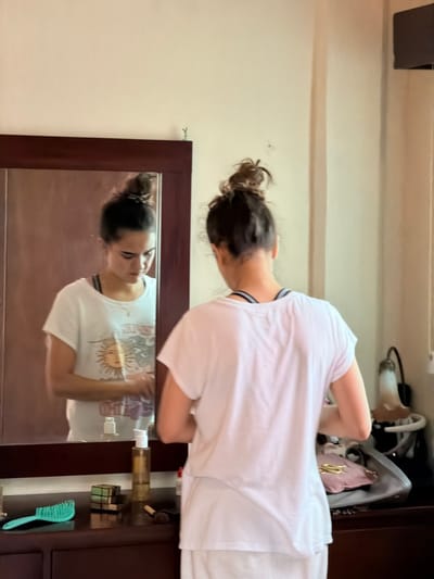 A woman with her hair in a bun stands in front of a mirror, her reflection visible. She wears a white t-shirt, different from the one in the mirror. The reflection's shirt features a sun graphic. A dark wood vanity is behind her, holding various items: bags, toiletry bottles, a Rubik's cube, and a teal hairbrush. The background includes a plain wall with a dark fixture on the ceiling.