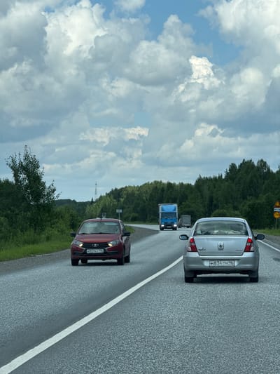 An aerial view from a vehicle captures a highway in Gornouralsky, Russia, under a cloudy sky. The asphalt road stretches ahead, with a white line dividing the lanes. A red sedan is visible in the foreground, its license plate reading "M909KP 196." Further down the road, a silver sedan follows, displaying the license plate "M834TM 96." In the distance, a blue truck and a smaller truck are traveling in the same direction. The sides of the road are lined with dense green trees and vegetation. A cell tower can be seen in the background, rising above the treeline. A yellow road sign with the number "70" indicates a speed limit.