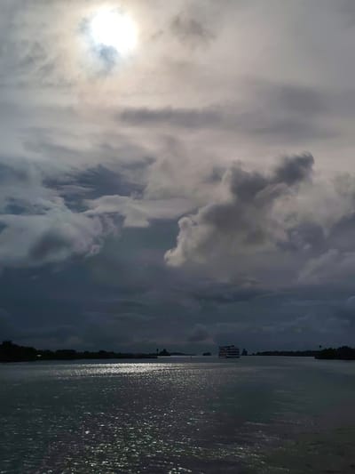 The image shows a wide, dark body of water, likely a river or bay, with a distant shoreline lined with trees. The water reflects a hazy, bright sun in the upper left of the frame, creating a shimmering effect. Dark, dramatic clouds fill the sky, with some lighter patches suggesting breaks in the overcast. A large, multi-decked boat is visible in the middle distance, moving across the water. The scene evokes a sense of twilight or an approaching storm. The location is identified as Kargulino, Russia.