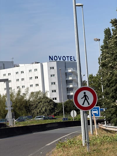 This media file shows a Novotel hotel building and its surroundings. The building is modern and white, with multiple floors and numerous windows. The Novotel logo is prominently displayed in blue letters on the side of the building.
In the foreground, a traffic sign with a red circle and a diagonal bar indicates that pedestrians are prohibited. Next to it, another sign shows a blue square with a white silhouette of a truck, likely indicating a truck route or parking area. A street lamp is visible above the signs, suggesting it is daytime. The scene includes trees and grass, with a road running alongside. There are cars parked on the side of the road. The sky is clear and blue.
The overall impression is a view of a hotel and its immediate vicinity, likely on a bright, sunny day. The presence of traffic signs suggests this is a public area with regulated access. The image appears to be taken in Rungis, France, as indicated by the location context. There is no explicit indication of specific emotions or activities taking place, other than the general context of a hotel and road infrastructure.