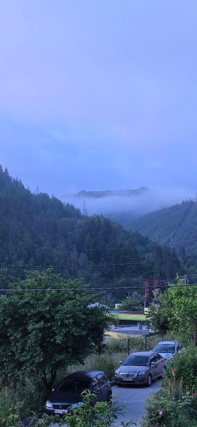 The image presents a serene, misty landscape, likely captured during early morning or late evening due to the soft, diffused light. The scene combines natural mountain scenery with elements of human presence.

On the left side of the frame, rolling hills are densely covered in evergreen forests. A thick layer of fog or mist partially obscures these forested slopes, particularly in the valleys and distant ridges, creating an atmospheric and somewhat ethereal backdrop. Several electricity pylons and power lines can be seen extending across the misty, wooded terrain. The sky above the hills is overcast and displays soft shades of light blue.

The right side of the image features a paved area or roadside, where three cars are parked. The closest vehicle is a dark-colored car, identified by its visible Bulgarian license plate "CM 075ABA". Behind it, a light brown or grey car, possibly a Toyota Avensis, also displays a Bulgarian license plate: "CM 0525 AX". A third, lighter-colored car is partially visible further back. Lush green vegetation, including trees and bushes, lines the roadside and surrounds the parked vehicles. A basketball hoop structure is also noticeable near the cars. In the mid-ground, beyond some trees and power lines, parts of a building with light-colored walls and a green roof are visible, alongside what appears to be a section of a sports field with white markings.

No specific activity is taking place; the scene is static, conveying a sense of quietude. The "CM" designation on the license plates confirms a Bulgarian registration, consistent with the location context of Rudozem, Bulgaria, nestled within a mountainous, forested region.