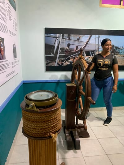 The image depicts a woman standing in what appears to be a museum exhibit in Boca del Río, Venezuela. She is dressed in a black t-shirt with a graphic and blue jeans, and is wearing black sneakers. The woman is standing next to a large wooden ship's wheel, with her right hand resting on it. To her left, a large brass compass is displayed on a wooden pedestal wrapped in thick rope. Behind the woman, a large photograph on the wall shows a boat under construction with two men working on it. The walls are painted in light blue and teal. The floor is tiled. The exhibit seems to focus on maritime history.