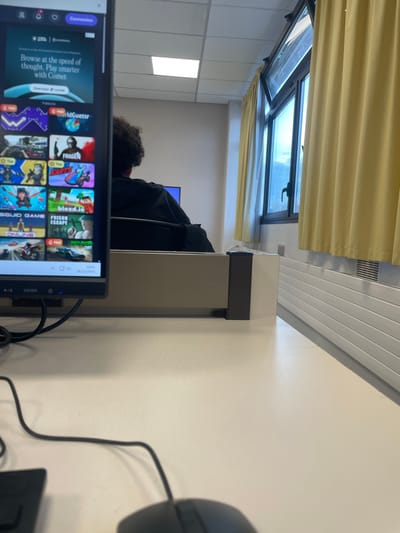 The scene depicts an interior view of a computer lab or classroom in Hérouville-Saint-Clair, France. In the foreground, a light-colored desk surface holds a black computer mouse and its cable. A vertically oriented black computer monitor, positioned on the left, displays a web browser or application launcher with a grid of game icons including "Worldguessr", "SQUID GAME", "bloxid.io", and "PRISON ESCAPE", along with an advertisement for "Comet". In the midground, the back of an individual with dark, curly hair, wearing a black garment, is visible, seated at a workstation behind a beige desk partition. To the far left background, a light-colored wall and ceiling with square fluorescent light fixtures are present, with a faint blue glow from another monitor. To the right background, a large multi-pane window, with its top section tilted inward, is flanked by drawn yellow curtains. Below the window, a white radiator with horizontal fins is mounted on the wall.