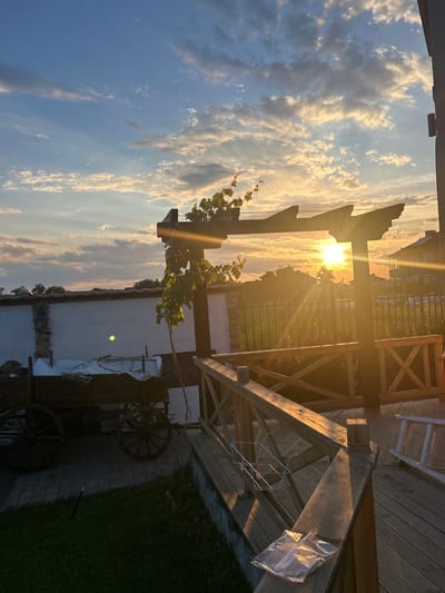 The image depicts a sunset over a rural property in Voyvodino, Bulgaria. The sky is filled with clouds, illuminated by the setting sun which casts long rays of light. In the foreground, a wooden deck with a railing is visible. A plastic bag rests on the railing. To the left, there is a white wall with a stone accent, and next to it, an old wooden cart with large wheels. A vine with orange flowers is growing on a wooden structure. Beyond the deck, a metal fence encloses a grassy area with some plants. In the background, houses and trees are silhouetted against the bright sky.