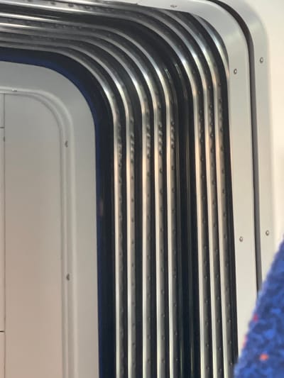 A close-up photograph shows the corner of an interior structure, likely a window or door frame, in Pardes Hana - Karkur, Israel. The frame is constructed of layered horizontal strips, alternating between light grey/silver and dark grey/black. The strips are curved at the corner. Small, circular fasteners (rivets or screws) are visible along the silver strips. A section of blue fabric is visible in the upper right corner. The overall impression is of a modern, possibly industrial, design element within a building or vehicle.