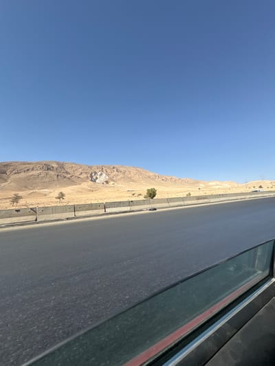 The image depicts an arid landscape in the City of Adra labor, Syria, viewed from inside a moving vehicle. A multi-lane asphalt road extends from the foreground into the midground, bordered on the left by a series of grey concrete Jersey barriers. Beyond the barriers, the terrain consists of dry, light brown earth with sparse, scrubby vegetation and a few small, dry trees scattered across the landscape. In the background, a range of rolling light brown mountains is visible under a clear, intensely blue sky. A prominent, large white or light grey geological formation or quarry is discernible on one of the central mountain slopes. A dark, indistinct object lies on the road shoulder next to the concrete barriers in the midground, and a white vehicle is faintly visible on the road in the far distance. No people or direct interactions are evident. The scene is illuminated by bright daylight.