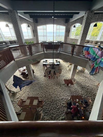 The image shows an indoor dining area with sand covering the floor, resembling a beach. The space has a mezzanine level with a wooden railing overlooking the main area. Large windows on the far wall offer a view of the ocean. Several tables and chairs are set up on the sand. In the foreground, a railing is visible. A large, colorful bird decoration is mounted on the wall to the right. On the mezzanine level, seating areas with sofas are arranged near the windows. In the main dining area, there are various wooden tables and chairs. A blue tarp is partially visible on the sand. Two people are seated at a table on the lower level, with one person in a maroon shirt leaning forward.