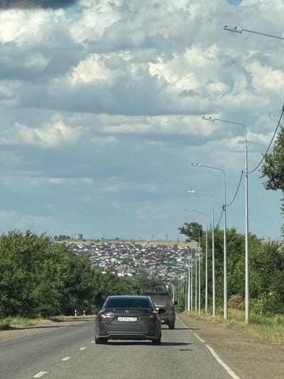 The media file shows a roadside view in Russia, likely during the daytime under a cloudy sky. The image is oriented vertically, with the road and a dense settlement of houses on the right side, and a large expanse of cloudy sky on the left.
The primary subjects are the road, a line of streetlights, a settlement of numerous houses with red and gray roofs, and a dark-colored car. The car, visible in the foreground on the right, has a license plate with the text "B819XC 09". The houses appear to be clustered together, forming a village or a suburban area. Green trees are present along the side of the road and also within the settlement.
The scene captures a snapshot of traffic on a road adjacent to a residential area. The activity taking place is implied to be driving or being a passenger in a vehicle. The lighting suggests it is daytime, possibly late afternoon or morning, given the presence of shadows and the overall brightness. The weather appears to be partly cloudy, with significant cloud cover in the sky.
Notable details include the repetitive pattern of streetlights along the road, the density of the housing settlement, and the license plate on the car. The image conveys a sense of everyday life in a Russian settlement with transportation infrastructure. There are no explicit emotional cues, but the scene depicts a common roadside view.