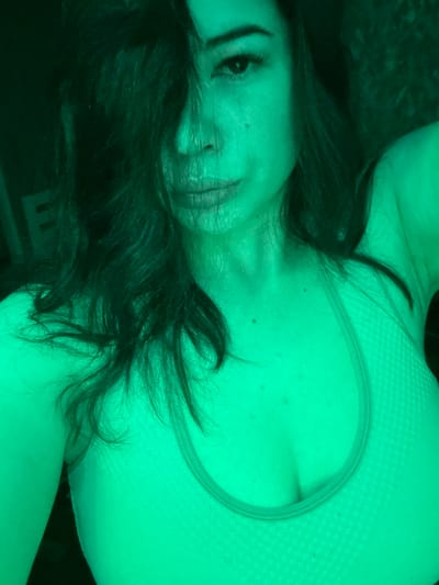 A close-up, low-angle, indoor, selfie-style photograph shows a woman's face and upper body. She has dark, wavy hair covering part of her face and is wearing a light-colored sports bra. The lighting in the image is a vibrant green, casting a strong hue over the entire scene. The woman's expression is neutral, with her lips slightly parted. She appears to be indoors, with a dark background that suggests a room. The city of Gavrilov Posad, Russia, is not depicted in this image.