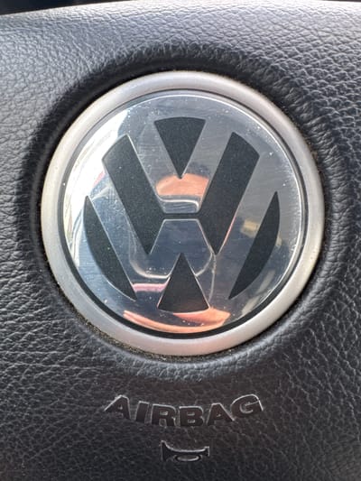 The image shows a close-up of a black leather steering wheel with the Volkswagen logo in the center. The logo is a silver circular emblem with the letters "V" and "W" superimposed on each other. Below the logo, the word "AIRBAG" is embossed in the leather. The steering wheel is textured and appears to be slightly worn. There is no indication of the location in Keila, Estonia.
