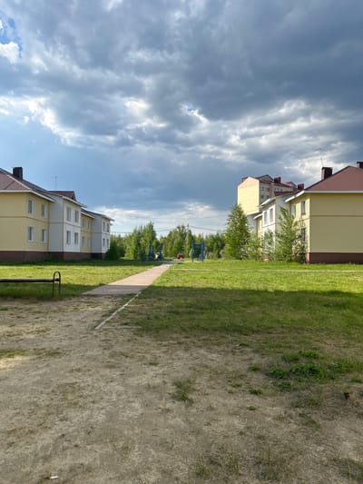 The image shows a residential area in Uray, Russia. Two-story buildings with yellow siding and dark red roofs face each other across a grassy area with a dirt path running between them. The sky is cloudy with patches of blue visible. In the distance, playground equipment is partially visible behind trees. There are no people visible in the image.