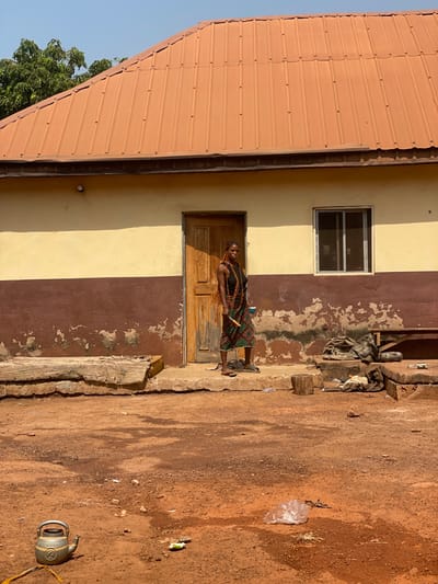 The image depicts a rural or semi-urban scene in Oturkpo, Nigeria, under bright sunlight. A single-story building occupies the middle ground, featuring a light yellow upper facade and a dark brown lower facade, with visible paint chipping on the lower section. An orange corrugated metal roof covers the structure. A light brown wooden door with vertical panels is centered on the building, to the left of which is a window with a silver frame and horizontal panes.
Standing on a concrete step in front of the door is a young adult female. She has dark skin and long, dark braided hair. She is wearing a dark, sleeveless top, a colorful patterned skirt (green, red, black, white geometric design), and dark sandals. Multiple strands of colorful beads adorn her neck and chest. She holds a light-colored cup in her left hand and a thin, light-colored stick or tool in her right hand, pointing downwards. Her gaze is directed towards the viewer.
To the left of the door, on the concrete step, two rough-hewn wooden logs are stacked horizontally. To the right of the door, also on the concrete, is a small wooden stump and various items including what appears to be a dark fabric sack, a metal basin, and debris, with a wooden bench partially visible behind them.
The foreground consists of reddish-brown, dry dirt ground with scattered small stones and patches of darker, damp-looking soil. In the lower-left foreground, a light grey metallic kettle, marked with the number "2," rests on the ground. In the lower-right foreground, a clear plastic bag and other small pieces of litter are visible on the dirt. Green foliage from a tree is visible in the upper-left background, behind the building. The sky is clear and light blue.