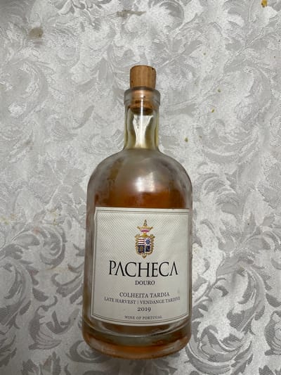 A clear glass bottle, partially filled with an amber-colored liquid, rests upright on a white, textured tablecloth. The bottle is sealed with a natural cork stopper and appears to have residue or condensation on its exterior surface. A white label on the front of the bottle displays a crest at the top, followed by the text "PACHECA DOURO", "COLHEITA TARDIA", "LATE HARVEST | VENDANGE TARDIVE", "2019", and "WINE OF PORTUGAL". The tablecloth features an embossed or woven floral/damask pattern and exhibits multiple dark brown and yellow stains, along with scattered small specks of debris. No people are present, and no specific actions or interactions are depicted. The exact location within the city of Mogadouro, Portugal, cannot be determined from the visual information provided in the image.