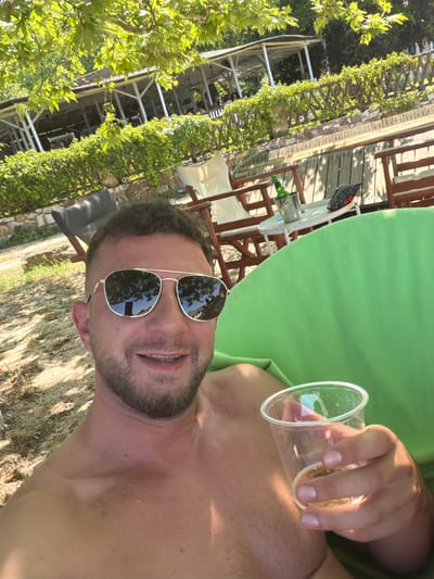 This detailed image, set in Ολυμπιάδα, Greece, depicts an outdoor scene featuring a single adult male. The subject, a Caucasian male with short dark hair and a beard, is positioned in the foreground, facing the camera with a slight smile. He is shirtless and wears aviator-style sunglasses that reflect overhead foliage and structures. His right hand holds a clear plastic cup containing a light-colored liquid. He is seated in a prominent, bright green, soft-sided chair. To the left of the subject, the ground consists of sand and sparse vegetation. In the mid-ground, a stone wall is visible, behind which rests a dark gray reclining beach chair. To the right, a small white circular table is situated between two wooden-framed beach chairs, one of which is partially obscured. On the table, a silver ice bucket holds a green bottle, and a black bag of snacks is present. The background features a low green hedge along a wooden fence, followed by a wooden deck. Beyond this, several white-painted wooden structures, resembling open-air pavilions or a restaurant, are visible amidst dense green trees and foliage, with faint figures of people discernible within the structures. The scene is brightly lit by natural sunlight.