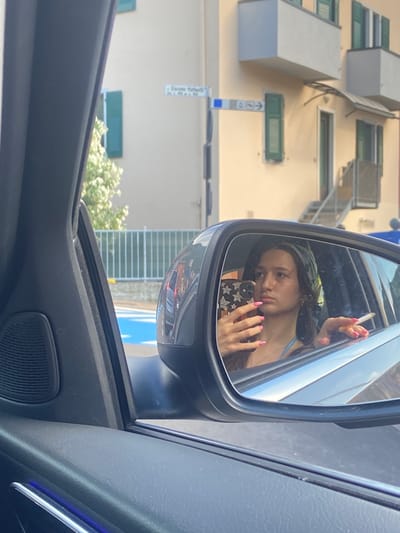 A young woman is captured in a reflection in a car's side mirror, holding a smartphone in her left hand and a lit cigarette in her right. She appears to be in a car parked on a street in Torbole, Italy, with a building and trees visible in the background. The building is a light yellow with green shutters and balconies. A street sign is visible on the building, reading "Giacomo Matteotti". The woman is wearing a blue bikini top and a green headscarf. Her nails are painted bright pink. The car's interior, including the door panel and speaker, is also visible in the foreground. The overall lighting suggests it is daytime.