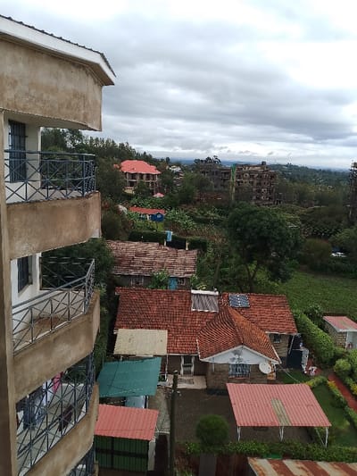 The image depicts an elevated view of a residential area in Olkeri ward, Kenya, under a uniformly overcast sky. On the left, the light brown concrete facade of a multi-story building occupies the foreground, featuring multiple balconies with dark grey metal railings exhibiting a geometric, cross-hatch pattern. Laundry is visible hanging on the railing of a lower balcony.

The midground reveals a landscape densely populated with various residential structures. Numerous houses feature terracotta tile roofs in shades of orange and red, with some equipped with solar water heaters and small photovoltaic panels. One house displays a white satellite dish. Interspersed among these are smaller structures with corrugated metal roofs in green, red, and off-white hues. A utility pole stands prominently. Extensive green vegetation, comprising trees and shrubs, fills the spaces between buildings and extends into the background, where a cultivated green field is visible.

In the background, the scene transitions to a more distant urban landscape. A multi-story building under construction, characterized by its exposed concrete framework, is discernible. Another building with a red tiled roof, potentially also under development, is present. A blue single-story structure is situated within a green clearing. The horizon consists of undulating green terrain beneath the cloudy sky. No individuals are visible in the image. A white vehicle is partially obscured by trees in the distance.