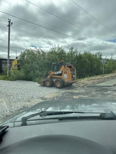 The image shows a construction site in Parma, Russia. A yellow CASE skid steer loader is parked on a gravel surface, surrounded by lush green trees. In the background, utility poles with power lines stretch across a cloudy sky. A sign with Russian text is visible on the left side of the image, indicating a rental service for construction equipment. The weather appears overcast, suggesting it might be a cloudy day. The scene is typical of an outdoor construction or landscaping project.