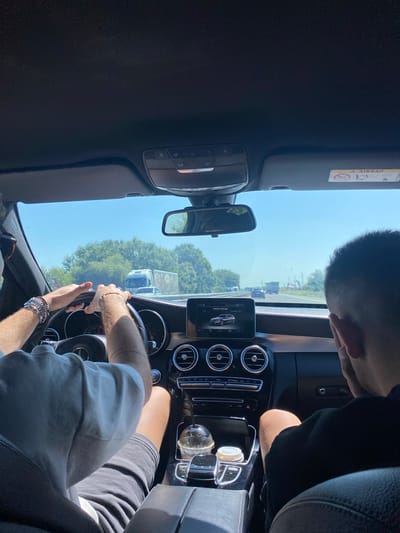 Two individuals are inside a Mercedes-Benz C-Class on a highway. The driver, on the left, is wearing a grey t-shirt and multiple bracelets on their left wrist. Their hands are on the steering wheel. The passenger, on the right, is wearing a black t-shirt and has their right hand near their face. The car's infotainment screen displays "Vehicle settings" with a graphic of a car and the text "DYNAMIC SELE." Outside the car, trees line the highway, and other vehicles, including a truck, are visible. The sky is clear and blue. The location is the A1 highway near Benkovski, Bulgaria.