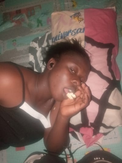 A woman reclines on a bed with patterned sheets and pillows. She is dark-skinned and wearing a black tank top with a white accent. Her hair is dark and appears tousled. She holds a piece of food in her hand, partially inserted into her mouth. She has dark, serious eyes and seems to be looking directly at the camera. Headphones or earbuds appear to be in her ears and a baseball cap sits near her head. The lighting is somewhat dim, creating a relaxed indoor scene.