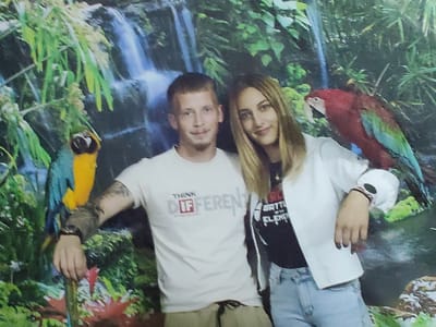 In Krushev Dol, Bulgaria, a young Caucasian couple poses for a photograph in front of a jungle-themed backdrop featuring a waterfall and two macaws. The male, on the left, wears a white t-shirt with the text "THINK DIFFERENT" printed on it, and khaki shorts. He has tattoos on his left arm and holds a stick-like object. The female, on the right, is dressed in a white jacket over a black t-shirt and light blue jeans. She wears a watch on her left wrist. The macaws, one blue and yellow and another red, are depicted in the background. The lighting is artificial and casts a soft glow on the subjects. The overall impression is a tourist snapshot taken in a studio or entertainment venue.
