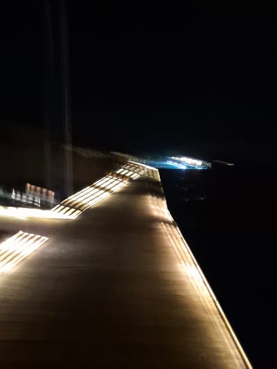 The image, taken at night in Bodufolhudhoo, Maldives, shows a building illuminated with artificial light. The building's facade is largely dark brown. The lighting creates strong shadows and highlights, emphasizing the texture of the wall. Some structural details like latticework are blurred due to motion. A sign on the left of the building shows white or brightly lit letters against a dark background. The sky is black. The overall composition is angled upward towards the building. There is no clear presence of people.