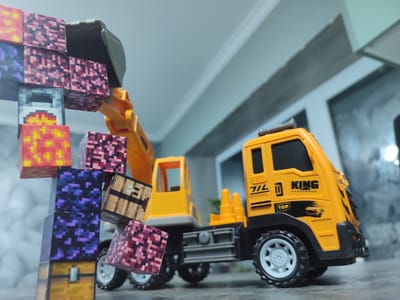 In Grigoriopol, Moldova, a close-up, low-angle shot features a toy excavator with a yellow cab and a black chassis, interacting with a stack of pixelated blocks resembling Minecraft game assets. The blocks, varying in color and pattern, are precariously stacked and appear to be in the process of being moved or loaded by the excavator's bucket. The background is softly blurred, showing a neutral, indoor environment with hints of walls and ceiling, suggesting the scene is set on a table or flat surface within a room. The lighting is diffused, casting subtle shadows and highlighting the textures of the toys.