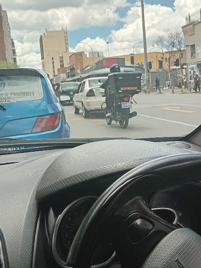 The image displays an urban street scene in Germiston, Gauteng Province, South Africa, captured from the interior of a vehicle. The immediate foreground shows a black, textured dashboard and the upper right portion of a black steering wheel, with a speedometer dial partially visible behind it indicating a low speed or RPM.
Directly ahead, in the midground, a light blue "Savvy" hatchback is observed, oriented away from the camera. Its rear window features a white decal with the text "PLUMBER" and "G & PROPERTY," a crest-like logo, and the partial numeric sequence "232." To the right of the hatchback, a person wearing a dark helmet and dark clothing is operating a black motorcycle. The motorcycle has a large black delivery box mounted on its rear, adorned with orange reflective tape. The motorcycle's license plate clearly displays "MH9 0MWGP." Directly in front of the motorcycle is a light-colored sedan, followed by a darker-colored sedan. The asphalt road surface visible includes yellow painted lane markings and directional arrows.
The background presents a typical urban environment with multiple multi-story buildings lining the street. A prominent, tall, light-colored building with a flat roof is situated on the left. Further down the street, an intersection is visible with traffic lights, one of which appears to be red. A street light pole is present on the right side of the road. Pedestrians are observable on the sidewalks to the right. The sky is partly cloudy with a mix of white clouds and blue sky.