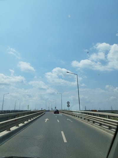 The image shows a view from a vehicle driving on a highway bridge. The sky is blue with scattered white clouds. The road is asphalt with white lane markings. There are metal guardrails on both sides of the road. Streetlights are visible along the bridge. In the distance, there are power lines and some buildings. A red car is visible in the right lane ahead. A speed limit sign indicating 50 km/h is visible on the right side of the road. The scene is set in the city of Olteni, Romania.