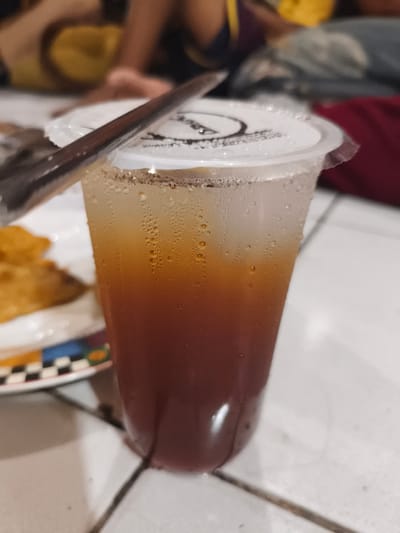 A transparent plastic cup containing an iced beverage with a gradient of dark reddish-brown liquid at the bottom transitioning to a lighter amber layer near the top is centrally positioned. The cup is covered in condensation droplets. A white plastic lid, sealed with a black circular logo/text, tops the cup, and a metallic spoon rests horizontally across it, partially covering the straw opening. To the lower left, a portion of a ceramic plate with a black and white checkered pattern and colored segments (blue, yellow, red, green) is visible, holding several blurry, golden-brown fried food items. The background, highly out of focus, shows multiple individuals in a casual indoor setting, including one person wearing a yellow top with a dark strap and another person in a red garment. The scene takes place on a light-colored tiled surface with dark grout lines. This image was captured in Cileungsi, Indonesia.
