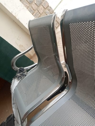 This is a close-up, high-angle shot of two perforated metal chairs, one grey and one black, positioned side-by-side. The grey chair, in the foreground, has a torn piece of fabric or padding attached to its left armrest. The chairs are situated outdoors, with a wall constructed of irregularly shaped stones and a section of a cream-colored structure in the background. A dark green band runs horizontally across the lower left portion of the image, suggesting a wall or barrier. The ground beneath the chairs appears to be a mix of dirt and possibly concrete. The scene is consistent with an outdoor seating area in a public space within Hotoro North, Nigeria, perhaps a waiting area or a park. There are no people or animals present in the image.