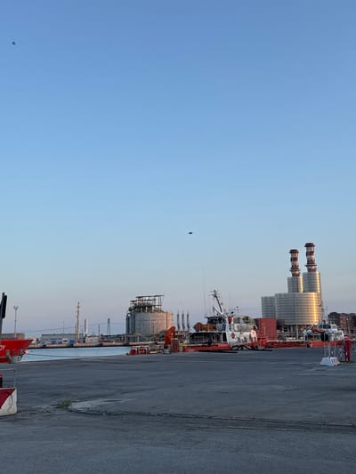 The image depicts an industrial port scene under a clear blue sky, likely in Ravenna, Italy, known for its port and industrial complexes. In the foreground, a paved concrete area leads towards the water. On the left, a large red vessel is docked, with the partial word "OY" visible on its side. Further into the scene, several boats are moored. Dominating the background are industrial structures: a large cylindrical storage tank with scaffolding on top, and two tall, distinctive smokestacks with red and white spiral patterns, characteristic of power plants. The water of the port is visible between the foreground and the industrial structures. Scattered throughout the sky are a few small, dark specks, which appear to be birds. The lighting suggests it is either early morning or late afternoon, with a warm glow illuminating the industrial elements.