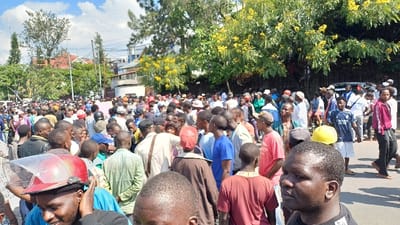 A large crowd of primarily adult males is gathered outdoors on a paved surface, likely a street, under a partly cloudy sky in Kabinzi, Rwanda. The individuals are standing closely together, facing various directions. Many are dressed in casual attire including t-shirts, shirts, and trousers; some wear hats or caps, and one individual in the foreground wears a red helmet with a clear visor. In the background, multiple buildings of varying sizes and styles are visible, interspersed with dense green foliage and trees featuring bright yellow flowers. Electrical poles and wires are also discernible. A white vehicle is partially visible on the right side, behind the crowd. The scene depicts a dense gathering of people in an urban or semi-urban environment.