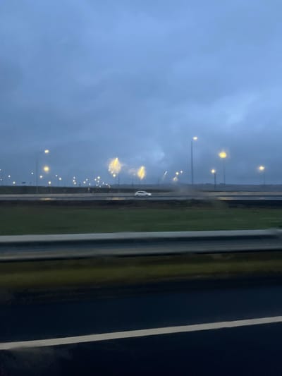 The image depicts a rainy highway at dusk, viewed from inside a vehicle. Streetlights line the road, reflecting in the wet pavement. A white car is visible in the distance. Dark clouds fill the sky above. The foreground shows parts of the vehicle's interior, including the window frame. The image is oriented in portrait format.