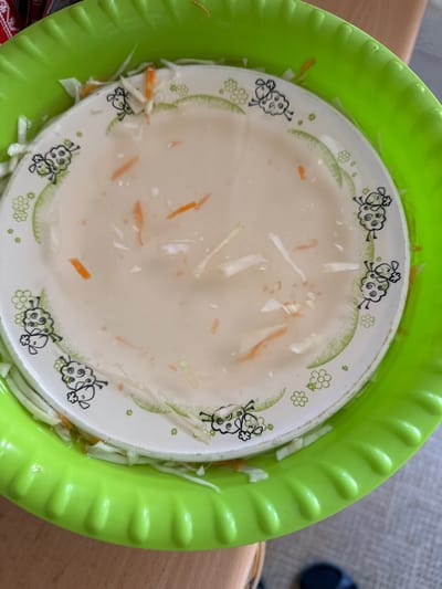 The image shows a top-down view of a white plate with a cartoon sheep design, placed inside a bright green plastic bowl. The plate contains a light-colored liquid and pieces of shredded vegetables, appearing to be cabbage and carrots. The vegetables are also visible around the rim of the plate, within the green bowl. The plate has a decorative border with small green flowers and sheep illustrations. The overall scene suggests a food preparation or presentation context. The lighting is natural, indicating it could be daytime. The background is blurred, showing a wooden surface and a hint of grey fabric, possibly a table and a chair or sofa. There is no visible text in the image.