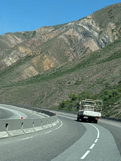 Here is a detailed description of the image:

The image shows a truck on a curved section of road. The road is marked with a solid white line and dashed white lines. A concrete barrier runs alongside the road. A steep, rocky mountain slope rises to the left of the road. The sky is blue. Based on the location provided, this scene is likely in the mountainous region near Krasnyy Most, Kyrgyzstan. The image captures a vehicle navigating a mountainous highway with safety barriers in place.