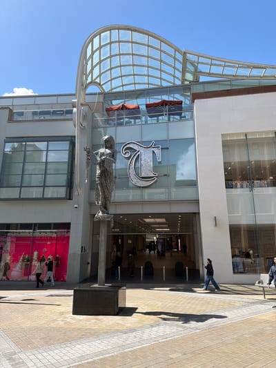 The image shows the Trinity Leeds shopping centre on a sunny day. A large, silver statue of a female figure stands in the foreground, to the left of the entrance. Above the entrance, a large, stylized letter "T" made of reflective materials is visible, along with the words "CENTRAL" and "LEEDS" written in reverse. The building features a curved glass roof and large windows. Several people are seen walking in the background, entering and exiting the shopping centre, and on the plaza. On the left side of the image, shop windows display mannequins and merchandise. The ground is paved with light-colored tiles.