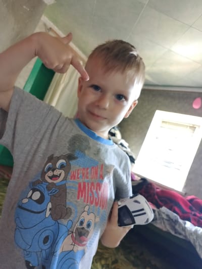 A young boy with blonde hair, wearing a gray t-shirt with a cartoon dog print and the words "WE'RE ON A MISSION", is looking at the camera and pointing towards himself. He is holding a small white toy car with black stripes in his right hand. The background appears to be an indoor setting with a window and some blankets. The image was taken in Pryvilne, Ukraine.