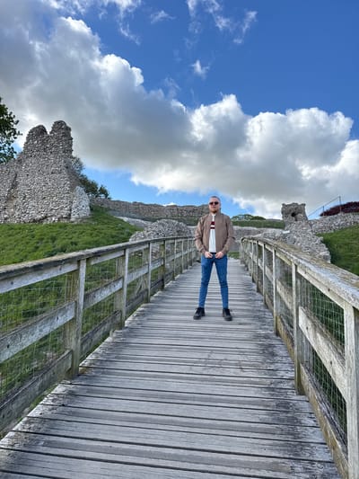 The image displays a man standing on a wooden walkway next to ancient stone ruins under a partly cloudy sky. The man is positioned centrally on the walkway, facing the viewer with his hands in his pockets. He is wearing a light brown jacket over a white and maroon striped shirt, blue jeans, dark shoes, and sunglasses. He has a beard and short hair.
The wooden walkway, constructed from planks, runs across the midground and foreground of the image, bordered by wooden railings with wire mesh. To the man's left and behind him are the weathered grey stone walls of what appears to be a historical ruin or castle, partially covered with green grass and some sparse vegetation. The background features a bright blue sky filled with large, fluffy white and grey cumulus clouds, suggesting a clear and sunny day. No text is visible in the image. The scene captures a relaxed moment at an outdoor historical site, possibly a popular tourist attraction.
