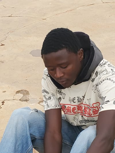 A young Black male with short, wavy black hair is seen from the chest up, leaning against a rough, cracked, light-colored wall. He is wearing a cream-colored t-shirt with a repeating pattern of black crosses and some red lettering. Over the t-shirt, he has on a dark grey hooded jacket, zipped up partially, with a black lining visible around the hood. The man's eyes are closed, and his head is tilted down, suggesting he may be sleeping or resting. His skin is dark, and his arms are visible, resting against his lap. He is wearing blue denim jeans, which appear slightly worn and have a distressed look, with a rip visible on the left knee.
The wall behind him is textured and shows signs of age and weathering, with prominent cracks and some peeling paint or plaster. The lighting suggests it is daytime, possibly outdoors, given the natural light and the texture of the wall. There are no obvious cues about the specific time of day or weather conditions beyond it being illuminated. The overall setting appears to be an outdoor environment in a somewhat rustic or older area. The emotion conveyed is one of peace or fatigue. The text on the shirt is in a stylized font, and the red lettering appears to be in English, but it is partially obscured and difficult to read completely. Some visible letters include "CHROME."