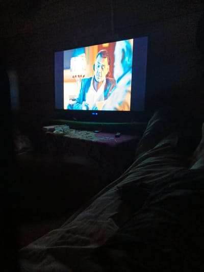 The image shows a television screen displaying a scene with two men. The man on the left is wearing a suit jacket over a white shirt, while the man on the right is wearing a light-colored shirt. They appear to be indoors, possibly in a room with dim lighting. In the foreground, there are blankets or bedding, suggesting someone is lying down while watching the television. The setting appears to be a domestic environment. There is no identifiable information in the image to determine the location in Strelka, Russia.