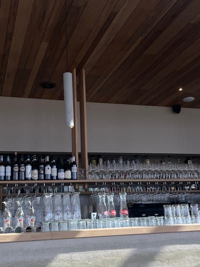 The image depicts the interior of a commercial establishment, likely a bar or restaurant, located in Overveen, Netherlands. The ceiling features dark brown wooden slats, from which a single long, light gray cylindrical pendant lamp with an illuminated base hangs. A black circular vent is visible on the left ceiling, and a recessed spotlight and a black security camera or speaker are on the right. Below the ceiling, the upper wall is a plain light cream or off-white. A multi-tiered bar structure, primarily wood, holds numerous items. The top shelf on the left contains multiple dark-colored wine bottles with various labels. The top shelf on the right displays an array of liquor bottles and rows of inverted stemmed wine or cocktail glasses. Lower shelves are densely populated with various types of clear glassware, including many inverted stemmed glasses, tumblers, and carafes bearing the "Stella Artois" logo. A dark commercial appliance, possibly a coffee or beverage machine, is visible behind the glassware on the lower right. The foreground features a light-colored, textured countertop with a smooth wooden edge. No individuals are present, and no actions or interactions are depicted.