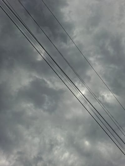The image captures a close-up, angled view of multiple parallel black electrical wires against a heavily overcast, gray sky. The wires are positioned diagonally from the bottom left to the upper right of the frame, with varying degrees of spacing between them. The sky is filled with dense, textured clouds, suggesting a gloomy or stormy weather condition. There are no people, structures, or other distinguishing features visible in the frame, making the wires and the sky the sole subjects of the photograph. The scene appears to be an outdoor environment, likely in an urban or semi-urban setting given the presence of electrical infrastructure. The specific location in Төмөнкү Ала-Арча, Kyrgyzstan is not visually discernible from the image itself, but the context implies the presence of such utility lines in that area.