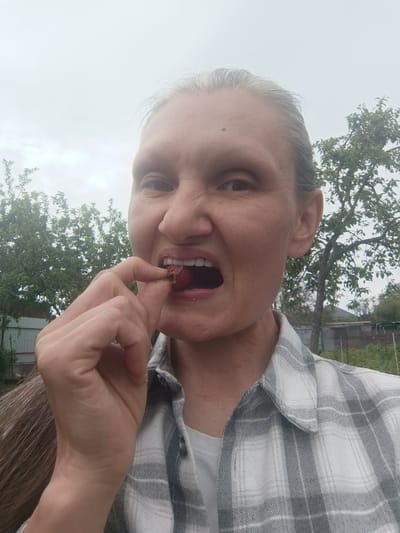 A woman with graying hair is taking a bite of a strawberry in an outdoor setting. She is wearing a gray and white plaid shirt and a white t-shirt underneath. The background consists of greenery from trees and bushes, a wooden fence, and a cloudy sky. The woman's face is close to the camera, and her mouth is open as she holds the strawberry to her lips with her fingers. The scene appears to be a garden or a rural area. The location is identified as Ostrogorka, Russia.