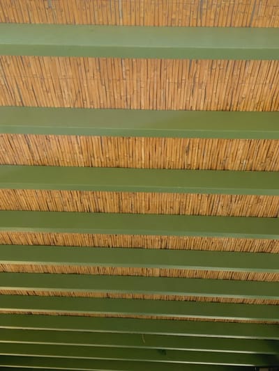 The image depicts the underside of a shaded ceiling structure located in Πλατύς Γιαλός, Greece. The structure consists of approximately twelve parallel, horizontal beams painted a uniform dark olive green. These beams are spaced evenly, with the gaps filled by tightly arranged, vertically oriented natural bamboo or reed poles, exhibiting various shades of light brown and tan. The arrangement forms a repeating pattern of green bands alternating with textured natural material. No individuals, specific objects beyond the structural components, or actions are discernible within the frame. The lighting is consistent across the visible surface.