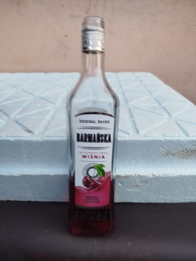 A clear glass bottle containing a dark red liquid with visible cherry pieces sits on a textured, light blue surface. The bottle has a silver cap and a label with the text "BARMASKA" in bold black letters. Below this, the label reads "WSZYSTKIE SKAR WYSOKIEJ JAKOŚCI" and "WIŚNIA", indicating a cherry-flavored product. The background consists of a pale, textured wall. The bottle and its contents are the central focus of the image. The scene appears to be indoors, possibly in Skórzewo, Poland, given the context.